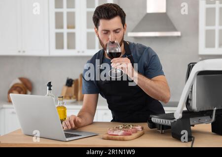 Mann mit einem Glas Wein beim Abendessen, während er sich den Online-Kochkurs mit einem Laptop in der Küche anschaute Stockfoto