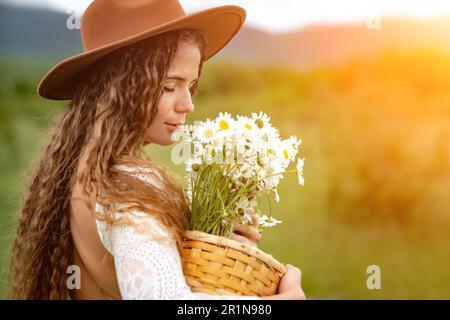 Eine Frau mittleren Alters in einem weißen Kleid und einem braunen Hut hält einen großen Strauß Gänseblümchen in den Händen. Wildflowers für Glückwünsche Stockfoto