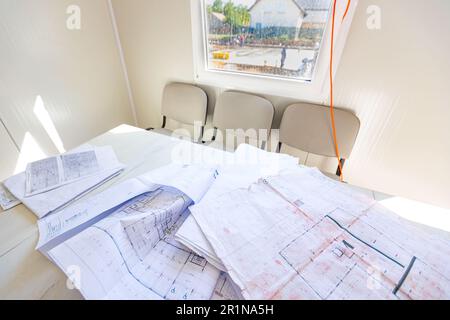 Masons Schreibtisch in einem Bürocontainer mit einem Haufen unordentlicher Architekturzeichnungen, Papierlayouts, Plänen. Fenster mit Blick auf die Baustelle Stockfoto