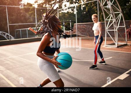Kann ich von hier aus ein Tor schießen? Eine vielfältige Gruppe von Sportlerinnen, die tagsüber gemeinsam ein Wettkampfspiel mit Basketball spielen. Stockfoto