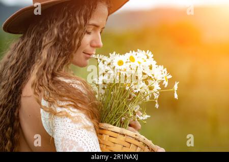 Eine Frau mittleren Alters in einem weißen Kleid und einem braunen Hut hält einen großen Strauß Gänseblümchen in den Händen. Wildflowers für Glückwünsche Stockfoto