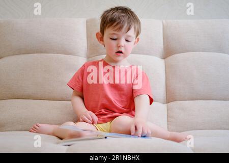 Das niedliche Baby spielt mit dem bunten Bilderbuch, blättert durch die Seiten und berührt die Bilder. Das kleine Kind liest Buch. Ein Kind im Alter von A Stockfoto