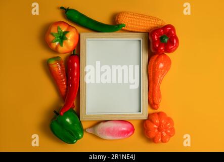 Leerer Bilderrahmen aus Holz mit Gemüse auf orangefarbenem Hintergrund, gesundes Essen im Hintergrund, vegetarisches Speisekonzept Stockfoto