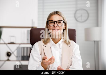 Lächelnde Geschäftsfrau mittleren Alters, die sich Webcam-Videokonferenzen ansieht, in einem virtuellen Chat-Meeting anruft und von dort aus einen Webinar-Kurs abhält Stockfoto