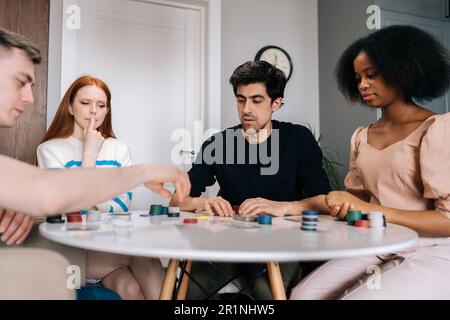 Nachdenkliche, junge, multiethnische Freunde, die am Wochenende eine Party feiern und Pokerkarten spielen, im Wohnzimmer sitzen und am Tisch sitzen. Stockfoto