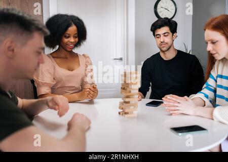 Eine multiethnische Gruppe fröhlicher Freunde, die ein Logikspiel spielen und zu Hause mit quadratischen Bauteilen und Würfeln gegeneinander antreten. Stockfoto