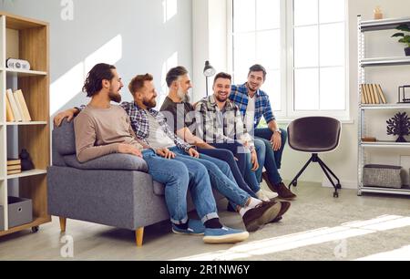 Männliche, bärtige Freunde, die zu Hause auf dem Sofa sitzen, lächeln und miteinander reden. Stockfoto