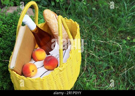Gelbe Korbtasche mit Buch, Pfirsichen, Baguette und Wein auf grünem Gras im Freien Stockfoto