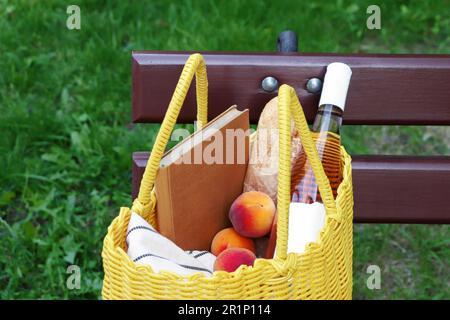 Gelbe Korbtasche mit Buch, Pfirsichen, Baguette und Wein auf Holzbank im Freien Stockfoto