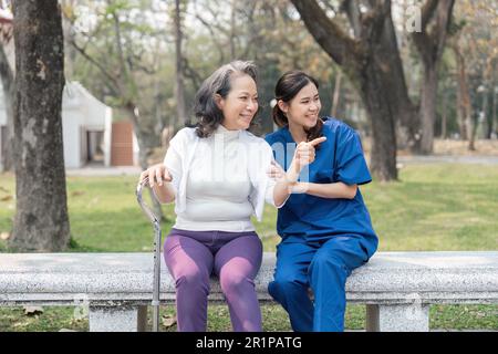 Eine asiatische Pflegekraft spricht, während sie mit einer alten Dame auf einer Bank in einem Krankenhauspark sitzt Stockfoto