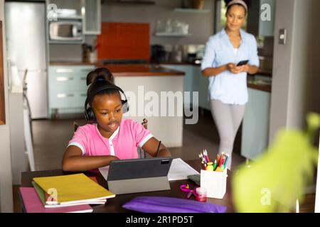 Glückliche afroamerikanische Mutter, die ihre Tochter mit Kopfhörern beobachtet, während sie zu Hause Online-Unterricht auf einem Tablet hat Stockfoto