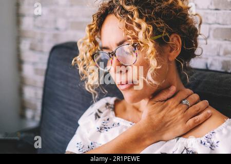 Kranke Erwachsene im mittleren Alter junge Frauen, die den Hals berühren, leiden unter Schulter- und Nackenschmerzen. Weibliche Schulterkranke zu Hause auf dem Sofa sitzen Stockfoto