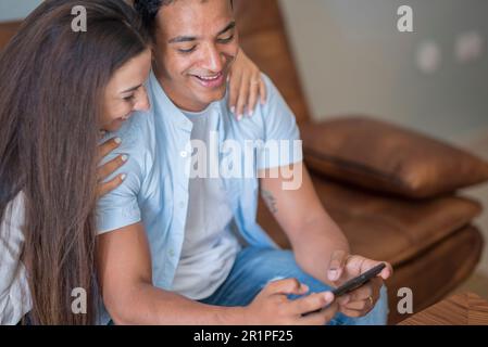 Junge glückliche Paare, Mobiltelefone teilen zu Hause Nachrichten aus sozialen Medien, lächelnder Ehemann und Ehefrau Millennials Kunden, die online einkaufen, sitzen am Tisch, Lifestyle-Konzept für mobile Technik Stockfoto