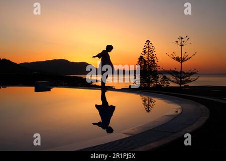 Junge Frau beim Sonnenaufgang am Pool, im Hintergrund am Strand in Kiotari, Rhodos, Griechenland Stockfoto