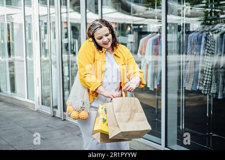 Fröhliche, übergroße, kurvige Frau, die draußen telefoniert. Außenporträt einer fröhlichen, lächelnden, aufrichtigen Frau in Übergröße mit Handy in der Stadt Stockfoto