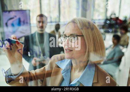 Selbstbewusste blonde, reife Geschäftsfrau in formeller Kleidung und Brille, die sich Notizen auf transparentem Brett macht und sie dem Publikum erklärt Stockfoto