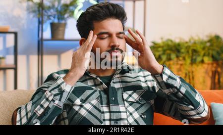 Verärgerter indischer Mann reibt Schläfen, um Kopfschmerzprobleme zu heilen, leidet an Spannung und Migräne, Stressgrimmen bei Schmerzen Bluthochdruck in der Wohnung zu Hause. Hindu-Typ auf der Couch. Lifestyles Stockfoto