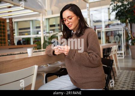 Mädchen benutzen Smartphone auf Gerät Bildschirm teilen soziale Medien Nachrichten tragen stylischen, trendigen Pullover Barhocker. Weibliche Smartphone-Nutzerin. Frau mit Brille, die ein Smartphone in der Hand hält Stockfoto