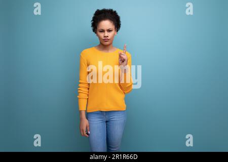 Junge lateinamerikanische Frau mit schwarzem lockigem Haar in einem gelben Pullover zeigt mit dem Zeigefinger nach oben Stockfoto