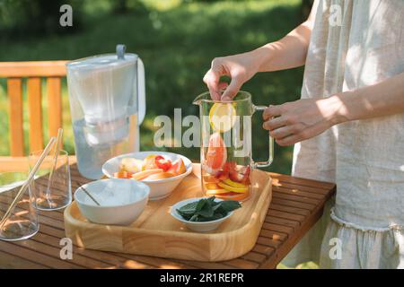 Frau bereitet im Garten eine Kanne Wasser mit verschiedenen frischen Früchten und Minzblättern zu Stockfoto