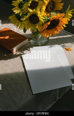 Ein Haufen Sonnenblumen in einer Vase, neben leeren Papierstücken und einem Hardback-Buch Stockfoto