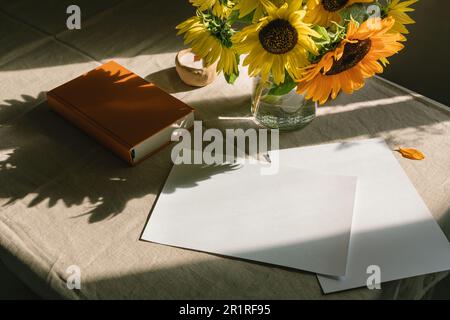 Ein Haufen Sonnenblumen in einer Vase, neben leeren Papierstücken und einem Hardback-Buch Stockfoto