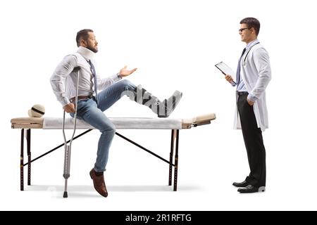 Verletzter Mann mit orthopädischem Stiefel und Halskragen, der auf einem Therapietisch sitzt und mit einem Arzt spricht, isoliert auf weißem Hintergrund Stockfoto