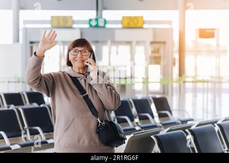 Eine glückliche, lächelnde, reife Frau hält ihr Smartphone und winkt jemandem zu, der sie am Flughafenterminal nach der Ankunft von der Reise um n abholt Stockfoto