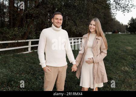 Ein junges, glückliches, romantisches schwangeres Paar geht an warmen Herbsttagen mit Händchen nach draußen. Schwangere Frau in Strickkleid und ihr gutaussehender Mann erwarten Stockfoto