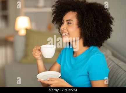 Lachende, fröhliche junge schwarze, lockige Frau trinkt eine Tasse heißen Kaffee auf dem Sofa, genießt Freizeit Stockfoto