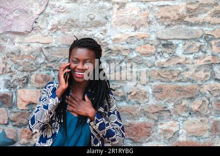 Selbstbewusste junge afrikanische Erwachsene Frau, die ein Handy hält, bekommt gute Nachrichten, lächelt, glücklich, draußen im Dorf über einer Mauer. Stockfoto