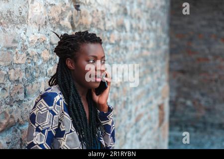 Selbstbewusste junge afrikanische Erwachsene Frau, die ein Handy hält, bekommt gute Nachrichten, lächelt, glücklich, draußen im Dorf über einer Mauer. Stockfoto