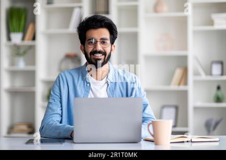 Attraktiver Junger Indischer Mann Sitzt Am Schreibtisch Mit Laptop Zu Hause Stockfoto
