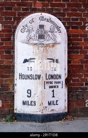 Nahaufnahme eines Meilensteins aus Metall vor einer roten Backsteinmauer in Westminster, London, England, mit der Entfernung von Kensington Road nach Hounslow Stockfoto