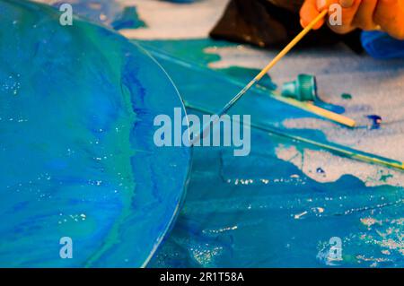 Der Prozess, ein hausgemachtes, modernes, abstraktes Muster zu kreieren, das mit einem Pinsel aus mehrfarbigem Acrylharz auf einem runden Holzbrett bemalt wurde. Stockfoto