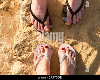 Zwei Paar männliche und weibliche Beine mit einer Maniküre in Pantoffeln, einem Fuß mit Fingern in Flip-Flops auf einem Sandboden aus Stein, Erde, Strand in einer tropischen wa Stockfoto