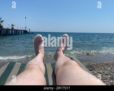 Männliche haarige Beine eines Urlaubers auf einer Sonnenliege am Strand am Meer. Stockfoto