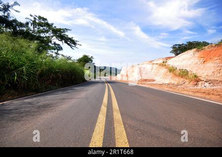 Geschwungene Straßenschilder mit horizontaler Beschilderung und seitlicher Hanglage. Blick von unten mit grüner Vegetation an der Seite und Himmel mit Wolken im Hintergrund Stockfoto