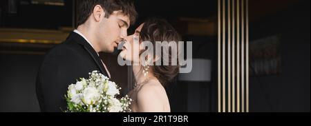 Seitenansicht eines gut aussehenden Bräutigams in Schwarz formell Kleidung küsst brünette Braut mit luxuriösem Schmuck hält Brautstrauß mit Blumen, während er steht Stockfoto