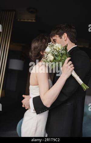 Braune Braut in elegantem Hochzeitskleid hält Brautstrauß mit Blumen und bedeckenden Gesichtern, während sie sich mit dem Bräutigam küsst, in schwarzer formeller Kleidung, während sie die Sta trägt Stockfoto
