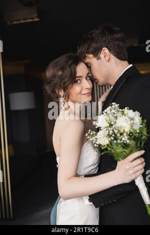 Brünette Braut in elegantem Hochzeitskleid mit Brautstrauß mit Blumen und umarmtem Bräutigam in schwarzer formeller Kleidung, während sie in der Hotellobby steht Stockfoto