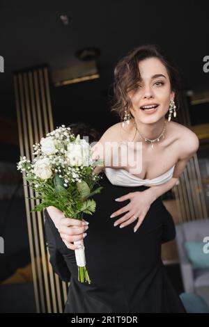 Bräutigam in schwarz formelle Kleidung Hebend hübsche Braut in weißem Hochzeitskleid und luxuriösen Schmuck halten Brautstrauß mit Blumen, während sie in heiß stehen Stockfoto