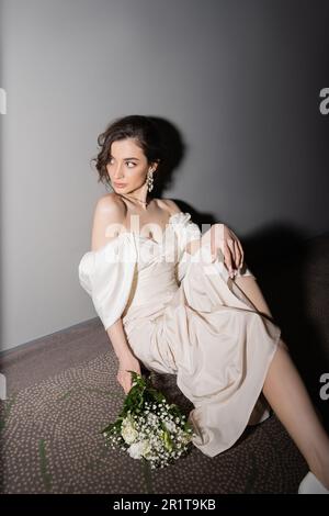 Strahlende junge Braut mit braunem Haar in weißem Hochzeitskleid, die wegschaut, während sie Brautstrauß mit Blumen hält und auf dem Flur in m sitzt Stockfoto