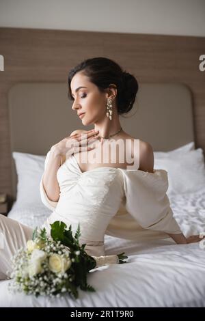 Bezaubernde junge Braut in weißem Kleid und luxuriösem Schmuck, die auf dem Bett neben dem Brautstrauß mit Blumen im modernen Schlafzimmer des Hotelzimmers sitzt Stockfoto