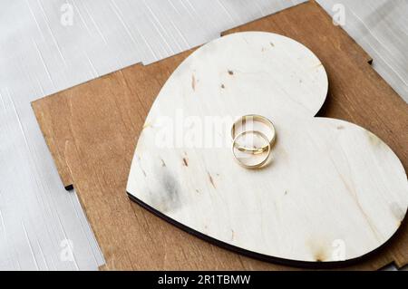 Goldene Eheringe auf einem hölzernen Herz. Leuchtende, glitzernde, glamouröse, modische, teure Herzen aus Holz mit Ornamenten zum Valentinstag aga Stockfoto