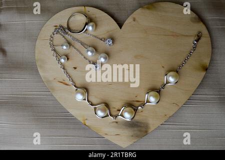 Ein hölzernes Herz zum Valentinstag mit silbernen Ornamenten, Ohrringen, Ringen, Halsketten mit Perlen, Diamanten, Edelsteine, Strasssteine auf einem beigen Ba Stockfoto