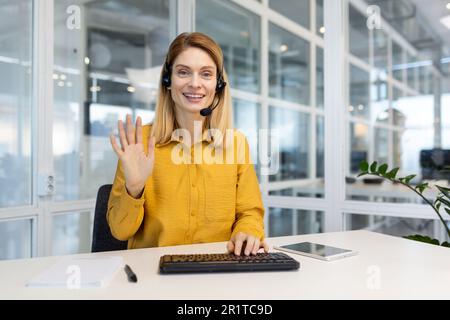 Porträt einer jungen Frau mit Headset, die im Büro an der Tastatur sitzt und die Kamera mit der Hand begrüßt. Arbeitet in den Bereichen Support und Assistance, Telemarket, Call Center. Stockfoto