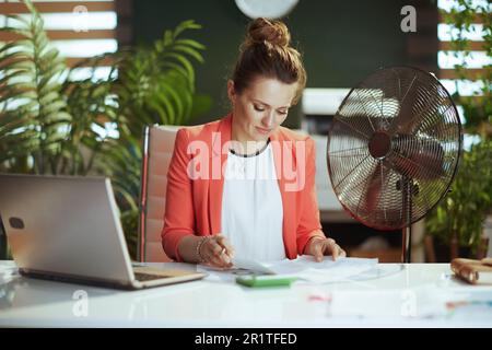 Nachhaltiger Arbeitsplatz. Moderne 40 Jahre alte Buchhalterin in modernem grünen Büro in roter Jacke mit elektrischem Ventilator und Laptop. Stockfoto