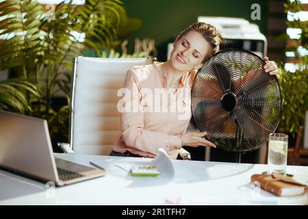 Nachhaltiger Arbeitsplatz. Glückliche, moderne Mitarbeiterin mittleren Alters in einem modernen grünen Büro mit elektrischem Ventilator und Laptop. Stockfoto