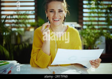 Glückliche, moderne, 40 Jahre alte Eigentümerin eines kleinen Unternehmens in einem gelben Pullover mit Dokumenten, die Videochats im modernen grünen Büro führen. Stockfoto